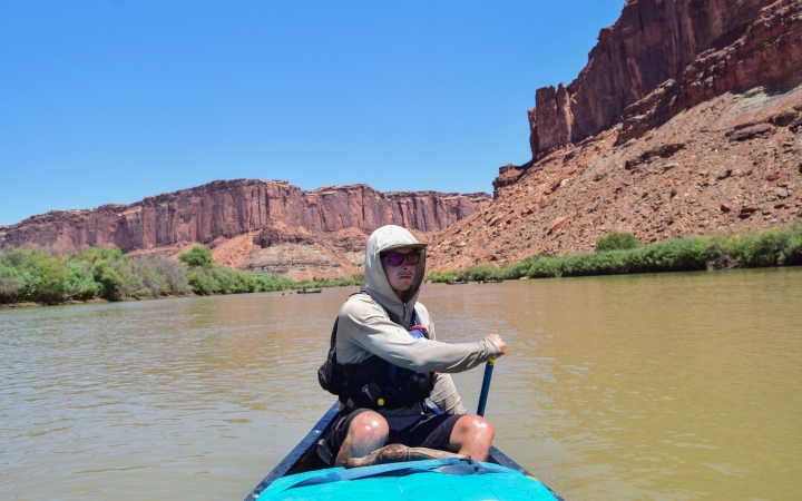 a person sitting at the back of a canoe paddles the watercraft forward. There are tall canyon walls behind them.
